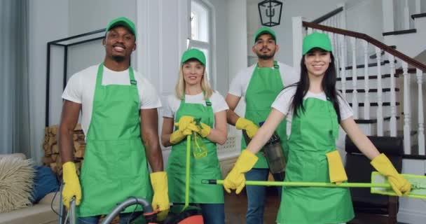 Portret atrakcyjnej uśmiechniętej wykwalifikowanej wielorasowej ekipy sprzątaczy w mundurach, która pozuje przed kamerą ze sprzętem do sprzątania w pięknym mieszkaniu — Wideo stockowe