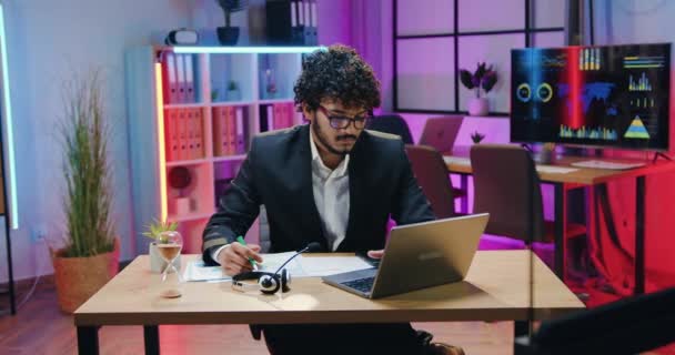 Attractive concentrated high-skilled bearded indian businessman working on laptop and writing out needed information into report in evening office — Vídeo de Stock