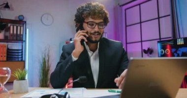 Güzel görünüşlü, başarılı, sakallı, resmi stil sahibi Asyalı iş adamı akşam ofisindeki dizüstü bilgisayarında çalışırken telefonla konuşmaktan zevk alıyor.