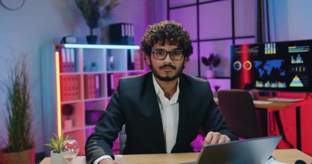 Attractive smiling successful skilled bearded muslim businessman in glasses sitting in front of camera in evening office with satisfied face expression,front view — 图库视频影像