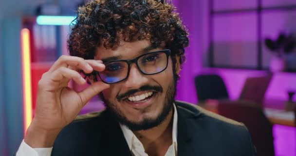 Крупним планом портрет гарного радісного успішного бородатого індійського офісного працівника в окулярах, який дивиться в камеру з щирою посмішкою в сучасному вечірньому офісі — стокове відео