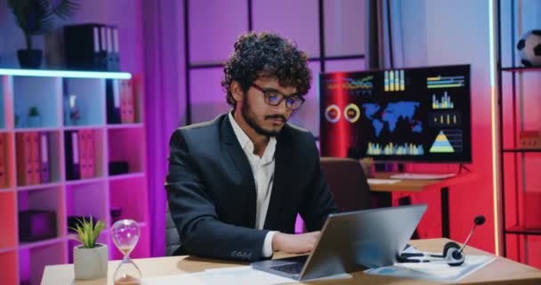 Ελκυστική θετική αυτοπεποίθηση έμπειρος γενειοφόρος μουσουλμάνος επιχειρηματίας σε γυαλιά που εργάζονται σε φορητό υπολογιστή και κοιτάζοντας στην κάμερα στο γραφείο το βράδυ, επιχειρηματική ιδέα — Αρχείο Βίντεο