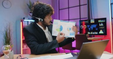 İş ortaklarının diyagramlarını bilgisayarda görüntülü sohbet yoluyla açıklayan kendine güvenen, kasıtlı, sakallı Asyalı iş adamının akşam ofisinde çalışmak.