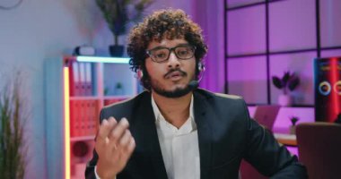 Sevimli, kendine güvenen, sakallı, kulaklıklı Hintli iş adamı akşam odası arka planında kameraya konuşuyor.