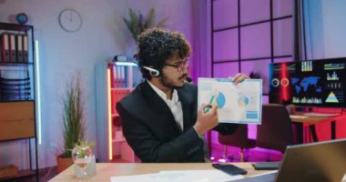 İyi görünümlü, kendine güvenen, kulaklıklı, modern, sakallı Arap iş adamı akşam ofisinde web kamerasının önünde oturuyor ve raporları diyagramlarla anlatıyor.