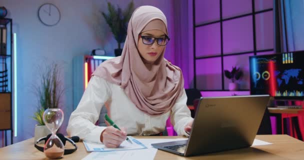 Pěkná sebevědomá pracovitá arabská podnikatelka v hidžábu kontrolující informace na obrazovce notebooku s daty na finančních zprávách ve večerní kanceláři — Stock video