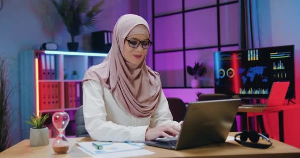 Atraktivní usmívající se sebevědomá profesionální arabská podnikatelka v šátku myšlení nad obchodní strategii a zadávání dat do počítače na pozadí večerní pracovní místnosti — Stock video
