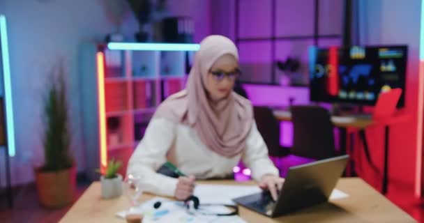 Telecamera ripresa su bella positivo fiducioso laborioso arabo ufficio donna managerin hijab che guardando in macchina fotografica mentre si lavora con documenti e laptop in ufficio serale — Video Stock
