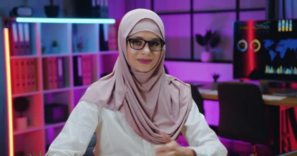 히잡 (hijab) 에서 자신감 있는 실력있는 아라비안 사업가가 저녁 사무실 배경에 엄지손가락을 들고 카메라를 보고 있는 멋진 미소짓는 모습의 초상화를 클로즈업해 보 세요. — 비디오
