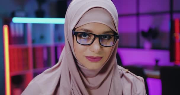 Retrato de muito sorrindo confiante de alta qualificada senhora de negócios árabe em luz violeta hijab em óculos que olhando para a câmera com expressão facial amigável na sala de trabalho à noite — Vídeo de Stock