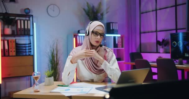 Remote work in office where good-looking confident motivated arabian businesswoman in hijab wears headset during online conference on laptop in the evening — Video Stock