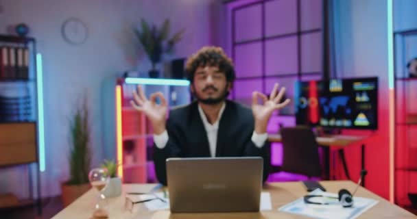 Camera shooting on handsome positive relaxed curly bearded muslim businessman in suit which sitting in front of camera with closed eyes and mudra hands in evening office — Vídeo de Stock