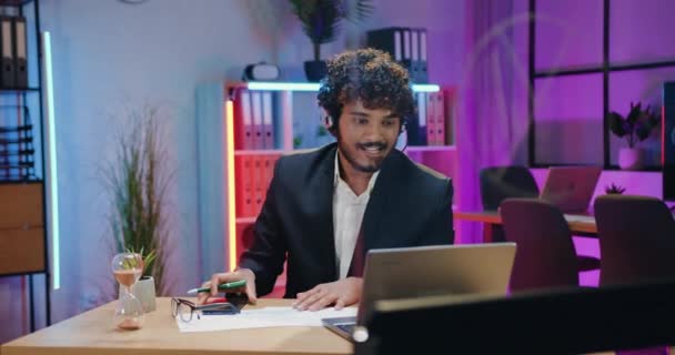 Attrayant positif confiant qualifié barbu homme d'affaires indien dans casque assis en face de l'ordinateur portable pendant le chat vidéo avec des partenaires d'affaires sur fond de bureau soir — Video