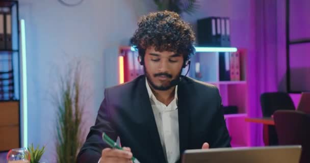 Çekici, neşeli, kalifiye, modern, sakallı, kablosuz kulaklıklı Müslüman ofis çalışanı iş ortaklarıyla çevrimiçi konferans sırasında bilgisayarın önünde oturuyor. — Stok video