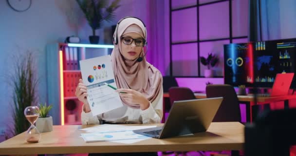 Zdalna koncepcja pracy, w której atrakcyjna odpowiedzialna, kompetentna arabska kobieta w hidżabie i zestaw słuchawkowy wyjaśniający raport z wynikami badań DNA podczas spotkania wideo na laptopie w wieczornym miejscu pracy — Wideo stockowe