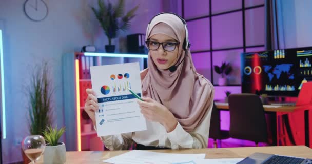 Krásná pozitivní sebevědomá zkušená arabka v hidžábu a ve sluchátkách sedí před kamerou ve večerní kanceláři a vysvětluje výsledky výzkumu DNA pro online diváky — Stock video