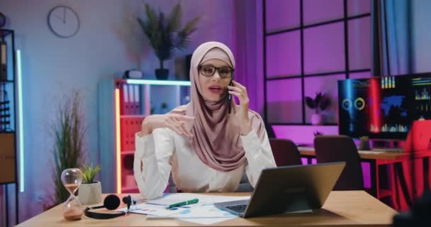 Lovely uśmiechnięty zadowolony arabski bizneswoman w świetle fioletowy hijab korzystających rozmowy telefonicznej w godzinach wieczornych biura podczas podejmowania pracy w godzinach nadliczbowych — Wideo stockowe