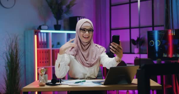 Atraktivní úsměv přátelská moderní arabská žena v hidžábu sedí na svém pracovišti ve večerní kanceláři během telefonního video chatu s přítelem nebo spolupracovníkem — Stock video