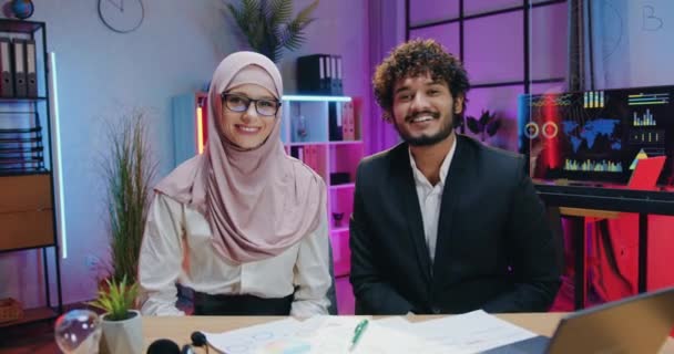 Good-looking smiling confident experienced muslim two businesspeople sitting in front of camera in evening office and gesturing thumbs up — Stock Video