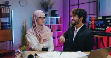 Mutlu gülümseyen başarılı Arap erkek ve kadın iş adamları akşam çalışma odasında kameranın önünde oturmuş birbirlerine beşlik çakıyorlar.