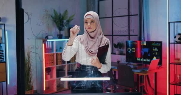 Zblízka pohled na krásné koncentrované kvalifikované tvrdě pracující arabské pracovnice v hidžábu, která stojí u skleněné stěny a dělá si na ní důležité poznámky ve večerní kanceláři — Stock video