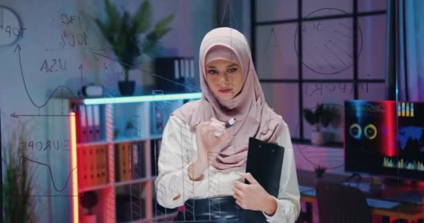View through glass wall on lovely serious creative high-skilled arabian female office manager in hijab which drawing on transparent wall with marker during brainstorming over project — Vídeo de Stock