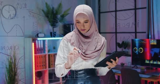 ガラスの壁の近くに立つヒジャーブの魅力的な集中熟練した勤勉なイスラム教徒の女性と夜のオフィスの壁にタブレットPCから重要なデータを書き出すビジネスコンセプト — ストック動画