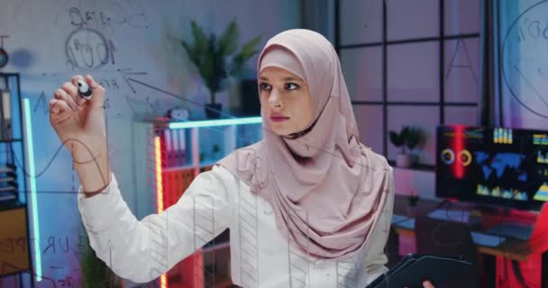 Potret wanita muslim terampil yang tampan tersenyum dan percaya diri dalam jilbab yang menulis di dinding kaca dan melihat ke kamera melalui dinding di latar belakang ruang kantor malam — Stok Video
