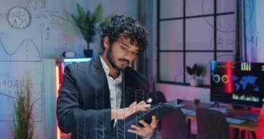 Yakışıklı, özgüvenli Hintli sakallı ofis çalışanı akşam ofisinde cam duvarın yanında duruyor ve bu duvardaki verileri tablet bilgisayardaki bilgilerle karşılaştırıyor.