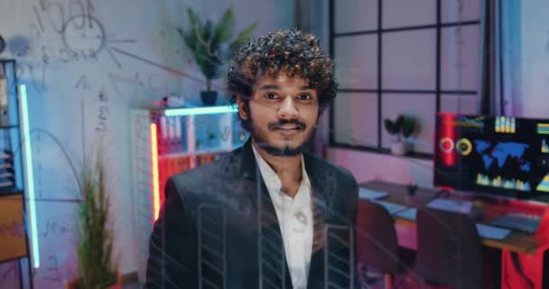 Aantrekkelijke glimlach succesvolle bebaarde moslim zakenman poseren op camera in het avondkantoor in de buurt van glazen muur tijdens het denken over zakelijke strategie van het project — Stockvideo