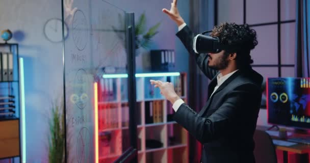 Atraktivní koncentrovaný moderní zkušený vousatý kancelářský manažer ve sluchátkách virtuální reality stojící před skleněnou stěnou ve večerní kanceláři a pracující na imaginární obrazovce — Stock video