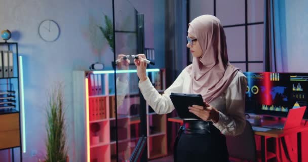 Belle concentrée arabe hautement qualifiée employée de bureau dans le hijab prendre des notes sur le panneau de verre lors de la navigation des informations sur tablette PC dans le bureau du soir — Video