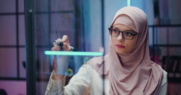 Camera shooting on pretty concentrated confident responsible young arabian businesswoman in traditional hijab which working on glass wall in evening office — Stock Video
