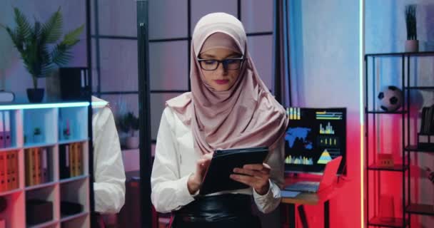 Primer plano retrato de atractiva sonriente confiada amigable experimentada musulmana mujer de negocios en hijab que los datos de navegación en la tableta PC y mirando a la cámara en la oficina de la noche — Vídeos de Stock