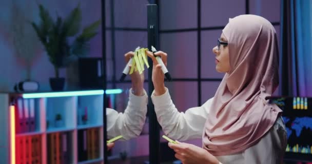 Side view of good-looking confident responsible hardworking skilled muslim female office worker in hijab which making importnat records on memo stickers and fixing them on glass wall — Stock Video