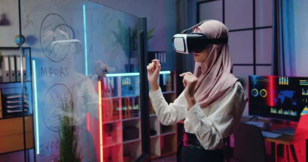 Σύγχρονη έννοια της τεχνολογίας, όπου ελκυστική συμπυκνωμένη έξυπνη, έμπειρη γυναίκα από την Αραβία με μαντίλα εργάζεται σε φανταστική οθόνη σε ακουστικά εικονικής πραγματικότητας στο βραδινό γραφείο — Αρχείο Βίντεο