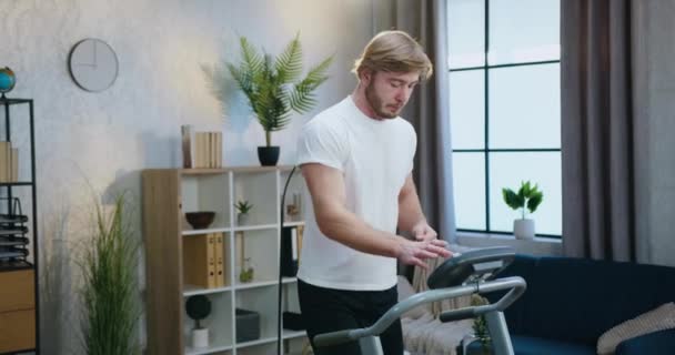 Aantrekkelijke sportieve jonge bebaarde man in sportkleding die tijdens de ochtendtraining thuis traint op hardloopbaan — Stockvideo