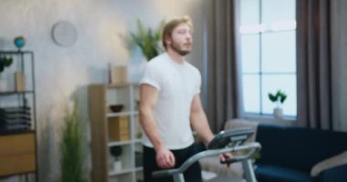 Yakışıklı, sportif sakallı, sportif bir adam sabah antrenmanı sırasında koşu bandında yürüyor ve dostça bir yüz ifadesiyle kameraya bakıyor.