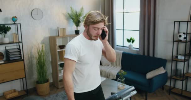 Beau gars barbu sportif positif en vêtements d'entraînement marchant sur la piste de course et parlant sur smartphone pendant l'entraînement à la maison, au ralenti — Video