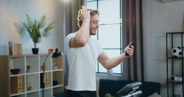 Polubiony młody brodaty mężczyzna w białej koszulce biegający na bieżni w domu i rozmawiający z przyjacielem przez telefon na czacie wideo — Wideo stockowe