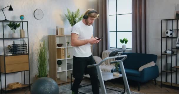 Home workout koncept där attraktiv aktiv sportig ung skäggig kille i sportkläder gör steppövningar på löpband och njuta av musik i hörlurar — Stockvideo