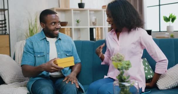 Atractiva alegre enigmática pareja afroamericana joven intercambiando cajas de regalo entre ellos, sentado tanto en el sofá suave en casa, concepto de emociones humanas — Vídeos de Stock