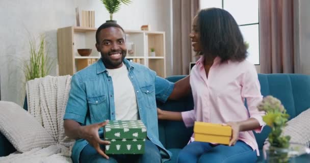 Snyggt glad leende kärleksfull afrikansk amerikansk par presentera presentförpackningar i kameran när du sitter tillsammans på soffan med kramar, framifrån — Stockvideo