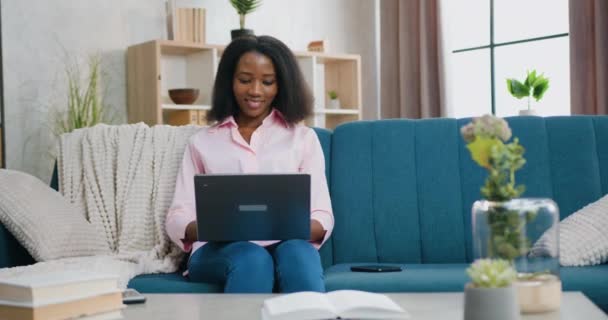 Linda feliz sorrindo despreocupado jovem mulher africana sentado no sofá macio em casa e sites de internet de navegação no computador, conceito de lazer — Vídeo de Stock