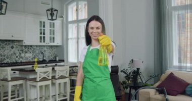 Eldivenli, gülümseyen, esmer bir kadın temizlikçi deterjan sıkar, kameranın önünde temizlik yapar.