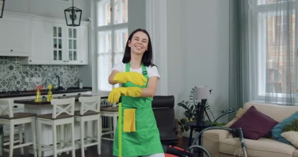 Sorriso atraente qualificado jovem morena trabalhadora da empresa de limpeza em uniforme posando na câmera durante a limpeza da casa do cliente — Vídeo de Stock