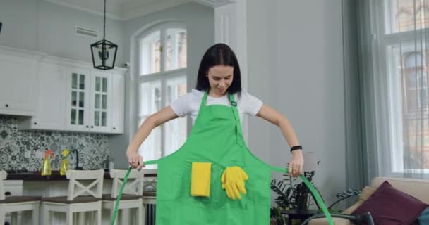 Piękny radosny młody wykwalifikowany kobieta sprzątaczka z usługi sprzątania wiąże fartuch przed rozpoczęciem pracy w domu klienta i patrząc na aparat z szczerym uśmiechem — Wideo stockowe