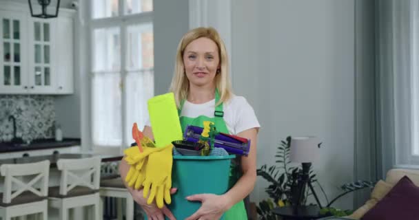 Pretty joyful skilled adult blond woman from cleaning service posing on camera with bucket with cleaning agents and equipment and showing gesture ok into camera during cleaning up customer flat — Stock Video