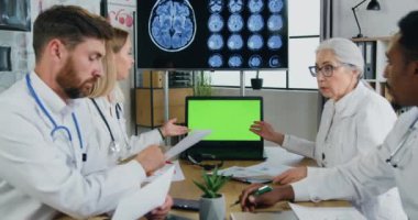 İyi görünümlü, kendine güvenen, çok ırklı tıp bilimcilerinden oluşan zeki bir ekip klinikteki ortak danışmanlık sırasında yeşil krom anahtar bilgisayar ekranı kullanıyor.