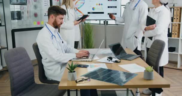 Handsome geconcentreerde gekwalificeerde bebaarde arts werkt op laptop, terwijl een andere mannelijke en vrouwelijke collega 's bespreken DNA structuur op whiteboard in zijn buurt in kliniek kantoor — Stockvideo